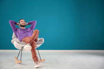 Handsome man listening to music while sitting in comfortable armchair against color wall