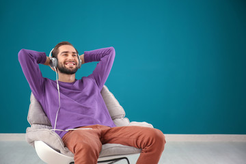 Handsome man listening to music while sitting in comfortable armchair against color wall