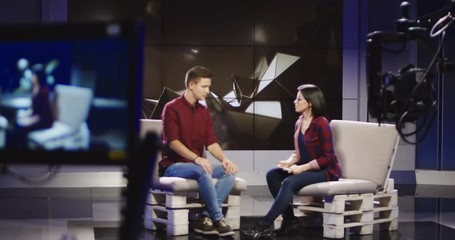 Young confident and relaxed man and woman taking participation in television show sitting in modern illuminated studio and having talk.