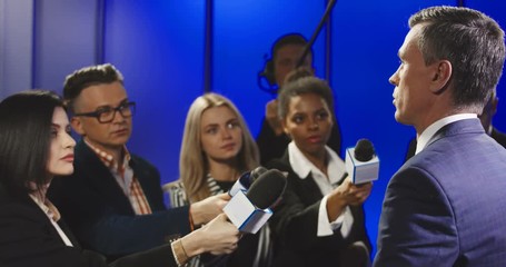 Big group of multiracial journalists with microphones and other technological equipment having interview with middle-aged politician in underlit studio.