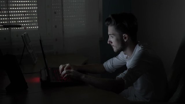 Excited male student sitting on his desk typing and chatting online with his girlfriend at night in a dark room
