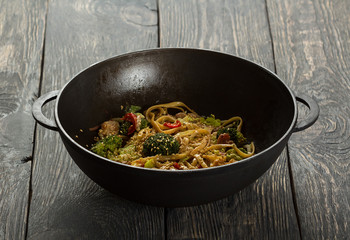 Spicy Asian noodles with meat and vegetables in wok, on wooden surface