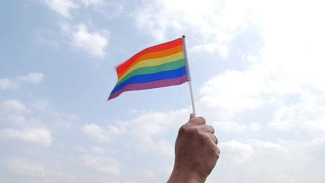 Hand holding LGBT flag against cloudy sky