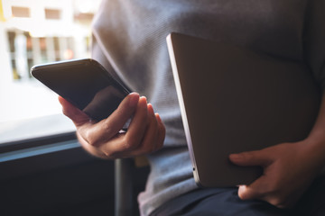Closeup image of a woman using and looking at a black smart phone while holding laptop computer