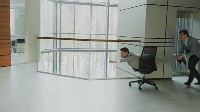 Slow motion pan shot of two funny businessmen riding office chair while having fun in lobby of modern business center