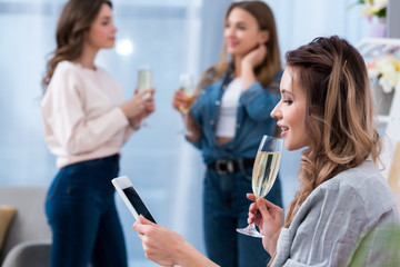 young woman holding glass of champagne and using smartphone while friends talking behind
