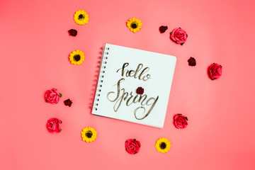 Notebook with white page with handlettering "Hello Spring" and various artificial flowers around. Flatlay, pink background.