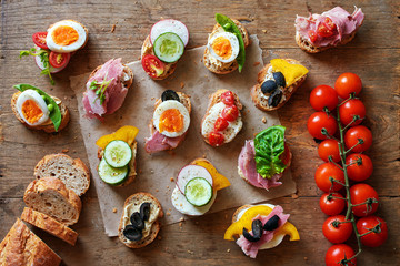 Top view of assorted sandwiches with baguette bread, cheese, ham, eggs, hummus and veggies. Healthy snack on wooden table.