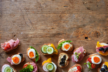 Variety of canape on baguette bread with ham and cheese, hummus, egg, tomato, olive and cucumber on wooden table with copy space. 