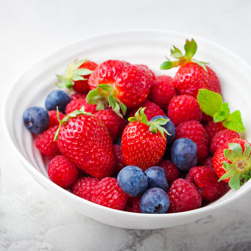 Fresh berries, blueberry, strawberry, raspberry with mint leaves in a white ceramic bowl on a gray stone background