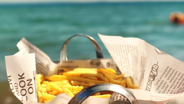 Close-up shot of woman hand taking appetizing golden crunchy French fries from the tray on sea background. Fast food snack ay the beach