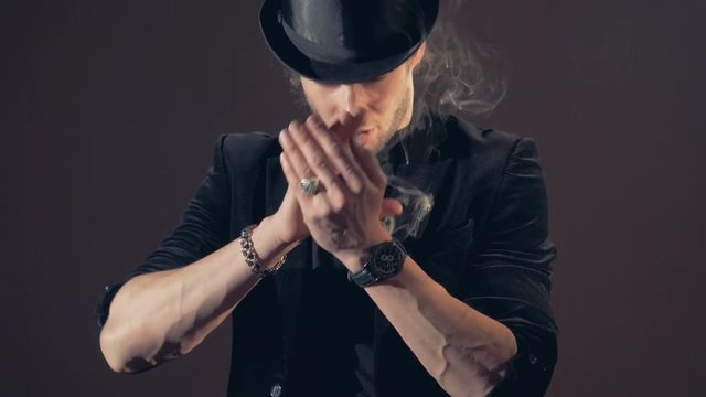 Magician is standing, his hands folded near his face, with smoke appearing.