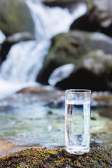 A transparent glass glass with drinking mountain water stands in the moss stone on sun beame against a background of a clean frost mountain river. The concept of drinking mountain drinking mineral