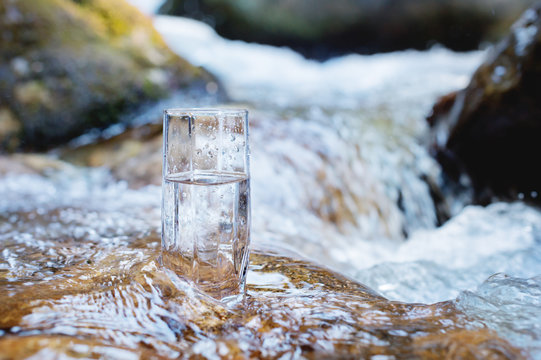 A glass glass with pure mountain drinking water stands on a rock in the course of a mountain river against the backdrop of seething cascades and waterfalls of a mountain river. The concept of pure