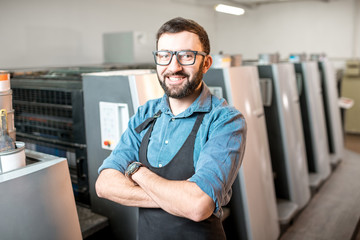 Portrait of a handsome typographer standing at the printing manufacturing with offset machine on...