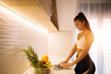 Young sporty girl using laptop computer on kitchen table while standing near window.