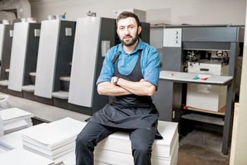 Portrait of a handsome typographer standing at the printing manufacturing with offset machine on...