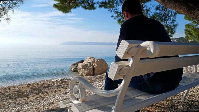 Young caucasian man sitting on wooden bench under pine tree on Adriatic coast and looking at sea