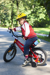 Fototapeta premium Little boy on bicycle in green park outdoor in summer. A child is riding a children's bike with support wheels wearing safety helmet