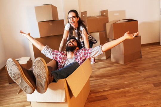 Young couple moving into a new home.Man sitting in cardboard box while woman pushes him all over the room.Real estate funny concept.