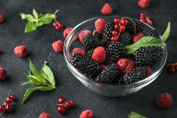 Glass plate with ripe red blackberries, raspberries, currant and green mint leaves on a black surface.