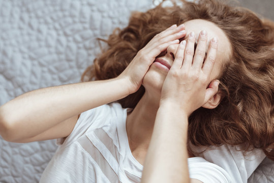 Need some rest. Top view on a depressed lady lying on a bed and covering her face with her hands while thinking about something negative.