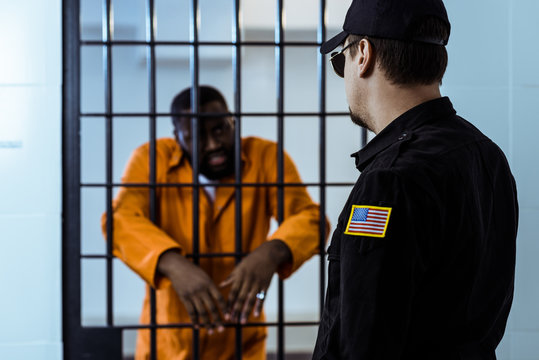 prison officer standing near prison bars and looking at african american prisoner