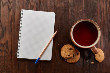 Cup of tea with cookies, workbook and a pencil on a wooden background, top view