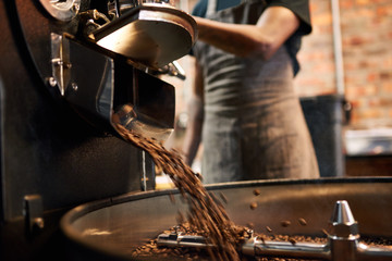 African man wearing an apron busy pouring coffee beans from the coffee roasting machine over to the tray that stirs the beans until they are cool enough to be packaged.
