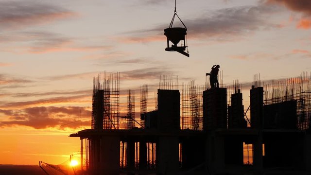 Silhouette of construction site agains sunset sky. Warkers wait when crane lifts concrete equipment