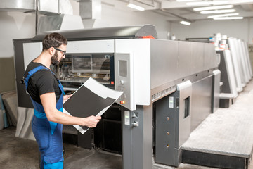 Typographer checking printing quality standing near the offset machine at the printing manufacturing