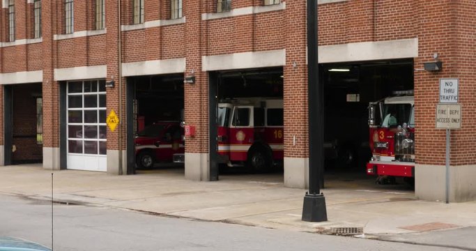 A daytime exterior establishing shot (DX) of a typical city red brick fire station with garage doors open and fire trucks parked inside.  