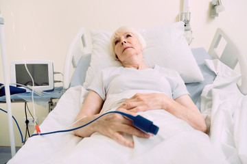 Obraz premium Getting better every day. Poor senior woman thinking about something pleasant while lying in a hospital bed with a heart rate medical equipment.