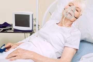 Severe illness. Sleeping senior lady lying in a hospital bed with a respiratory support and a heart rated medical equipment while undergoing treatment.