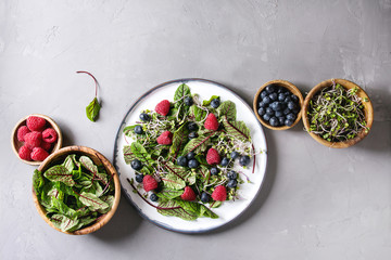 Fototapeta na wymiar Green vegan salad with berries strawberry, blueberry, sprouts, young beetroot leaves on ceramic plate. Ingredients in wooden bowls above over grey concrete background. Top view, space. Healthy eating