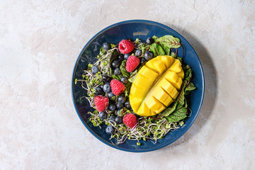 Green vegan salad with berries strawberry, blueberry, sprouts, young beetroot leaves, mango on blue plate over grey concrete background. Top view, space. Healthy eating