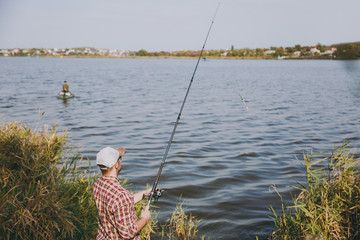 Back view Young unshaven man with fishing rod in checkered shirt, cap and sunglasses casts bait and fishing on lake from shore near shrubs and reeds. Lifestyle, recreation, fisherman leisure concept.