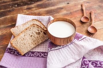 There are a wooden cup of milk, bread slices and two spoons on lilac dishcloth on diagonal old wooden planks.