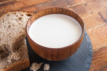 Wooden cup of milk and bread slices on kitchen round slate board on diagonal wooden planks background.