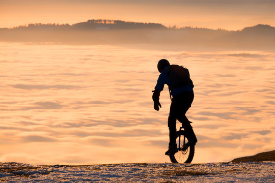 Man on unicycle riding on mountain over fog to sunset