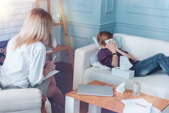 Unhappy memories. Teenage girl lying on a sofa getting emotional and wiping his eyes with napkins while talking with a professional therapist