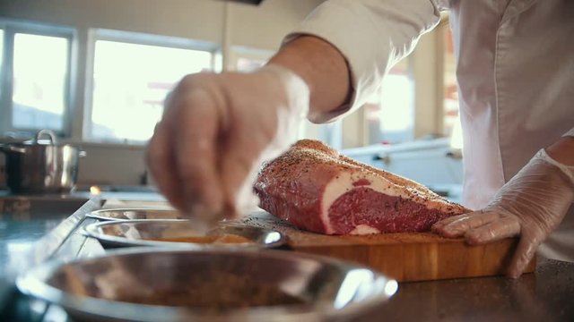 Butcher adding spices into large piece of fresh raw meat lying on a wooden board in a commercial kitchen, slow motion