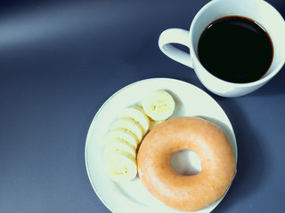 breakfast set from banana and donut with black coffee in white cup from morning time with blue background