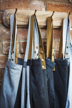 Denim work aprons hanging on a row of hooks on a brick wall.