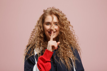 Stylish young girl student with curly hair shows a gesture quietly, secret. Curly hair. Pink background. Copyspace