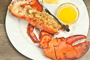 Close up, top view of, half, roasted lobster, stuffed with bread crumbs, sliced scallops on, round, white plate, steel nut cracker, artisan fork sliced lemon, glass bowl of melted butter