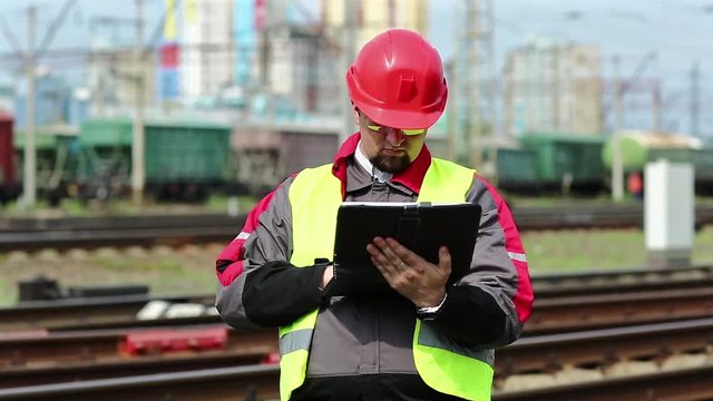 Railwayman with computer at freight train terminal.Railwayman in uniform and red hard hat works with tablet computer. Railway employee makes notes in his tablet pc