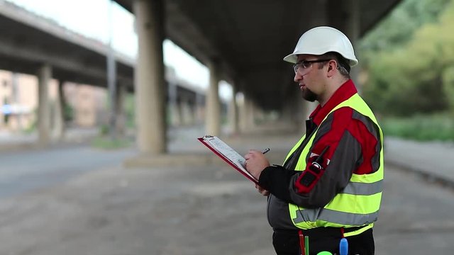 Road mender makes notes in work documentation. Building inspector with work documentation at project site under overpass