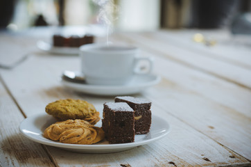 chocolate cookies and brownie in white plate on wood table with coffee cup at morning with sunrise. dessert and coffee concept.