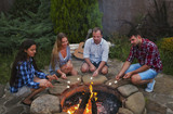 Company of young people holding sticks with marshmallows above campfire in the evening